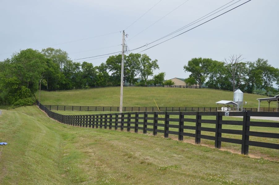 A black four-board farm fence curving along a sloping green pasture with a grain silo and barn behind