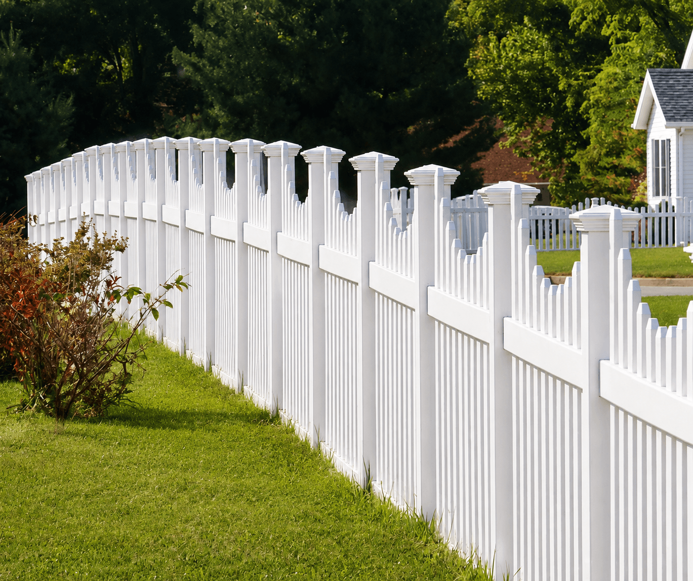 A white vinyl scalloped picket fence running along a landscaped suburban yard with a Sands Fence Co. sign