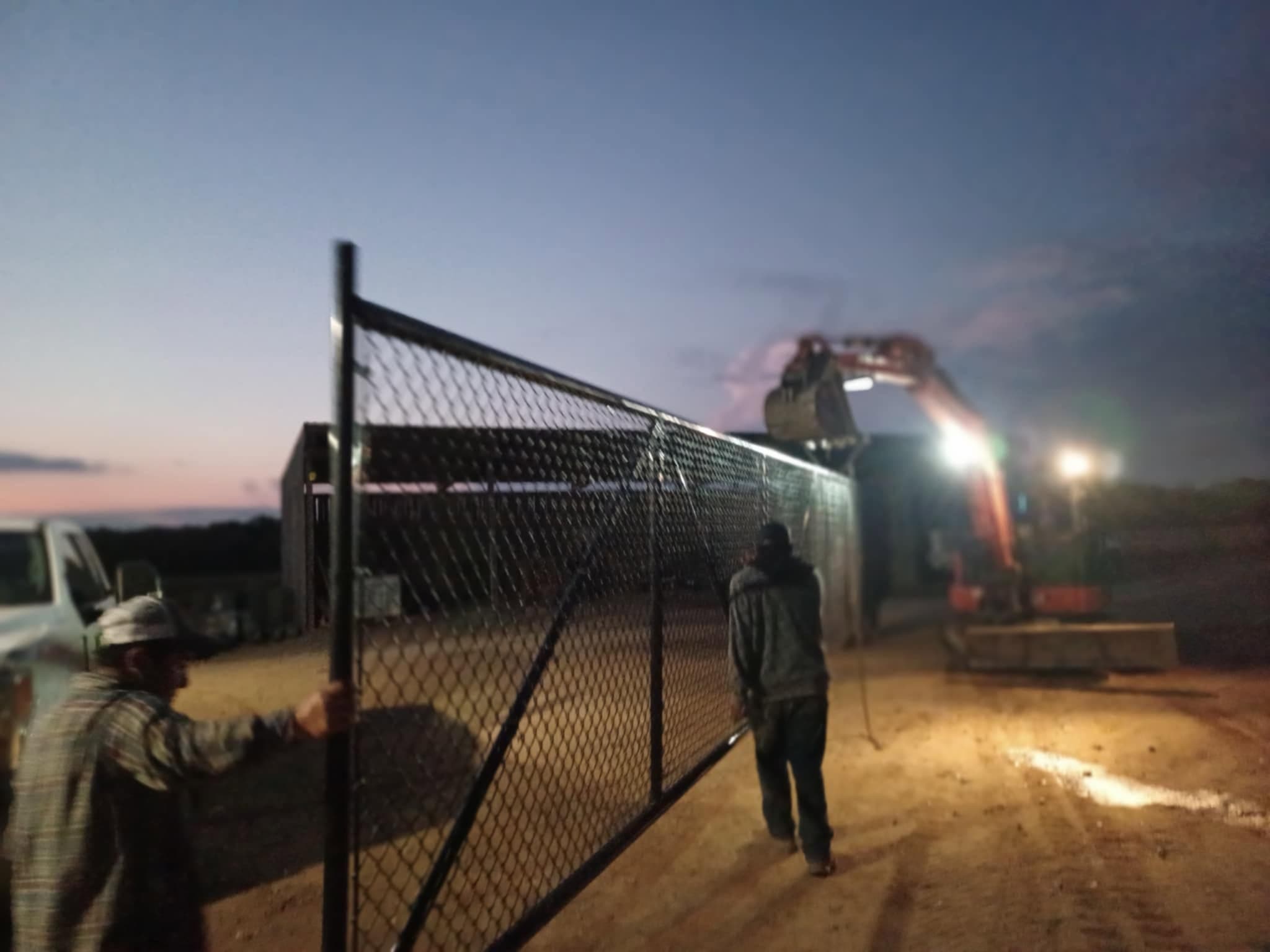 A Sands Fence Co. crew installing a black chain-link fence at dusk, lit by work lights