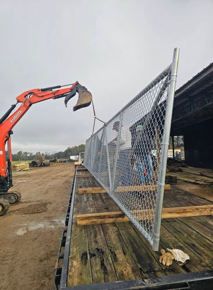 An excavator lifting a galvanized chain-link fence panel onto a flatbed trailer at the supply yard