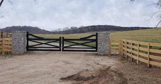 A black metal driveway gate between stone columns connecting to a wood farm fence