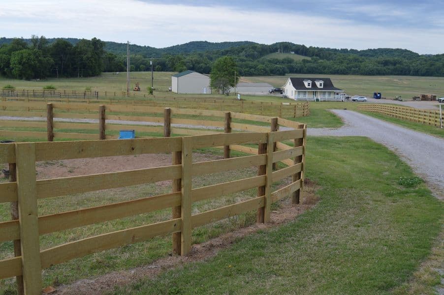 A four-board wood farm fence enclosing paddocks beside a gravel drive leading to a farmhouse, with rolling wooded hills behind
