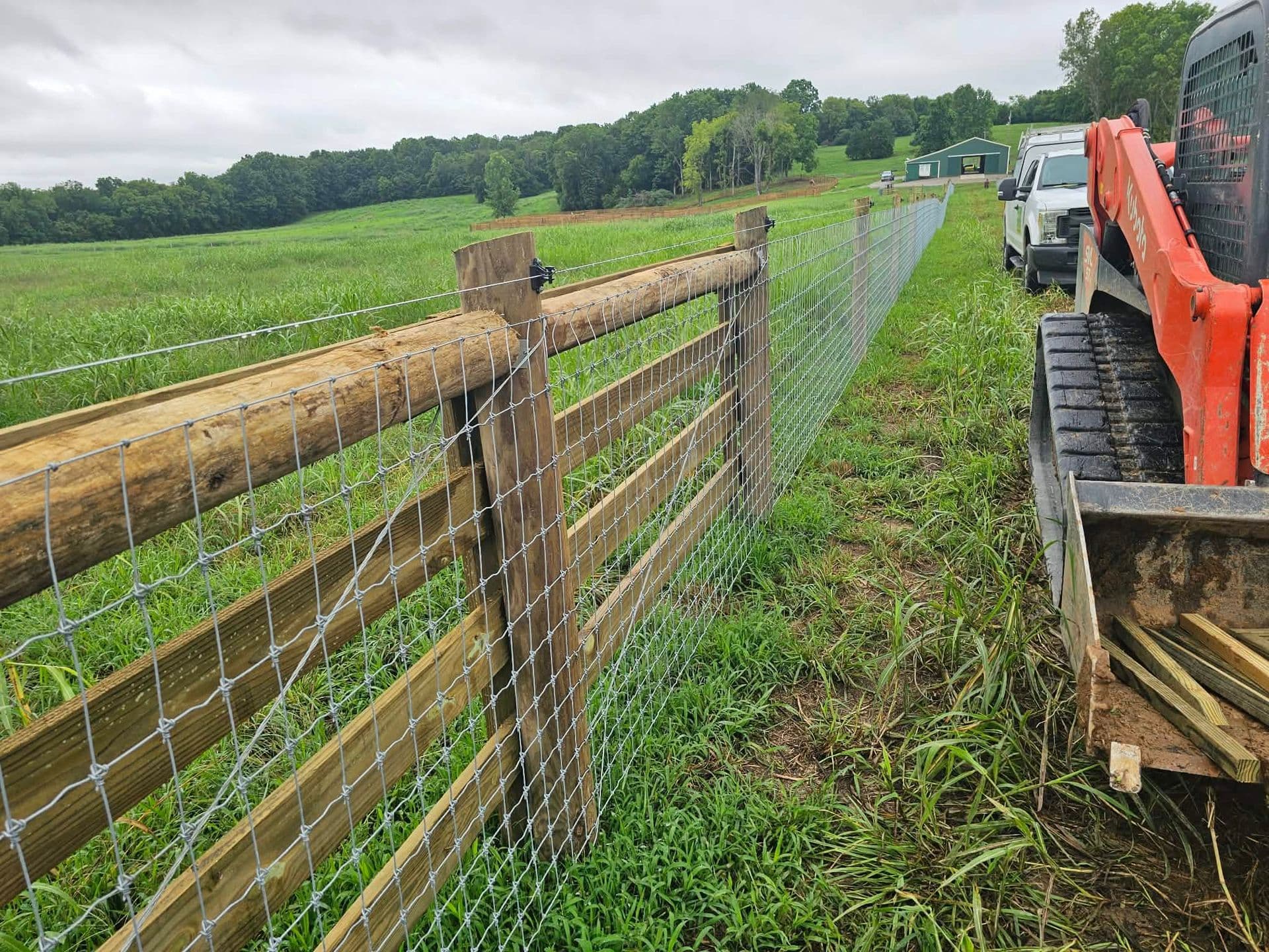 A combination wood-rail and woven-wire field fence running across a green pasture with a skid-steer and work trucks alongside