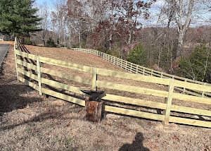 A freshly installed four-board wood fence running along a sloped rural property lined with autumn trees